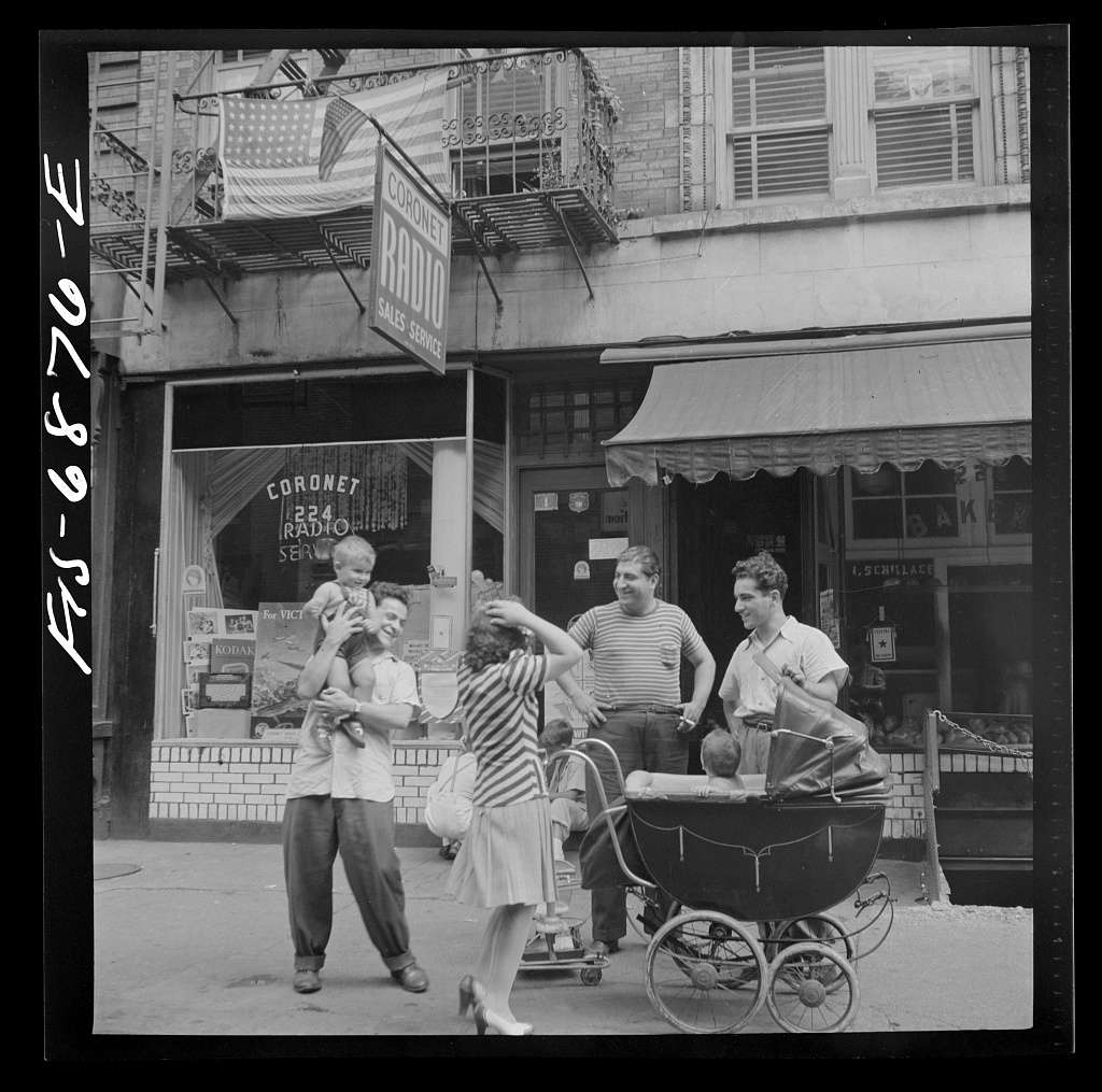 Adults and children gathered beside a baby carriage outside storefronts on Thompson Street, New York, in 1942.