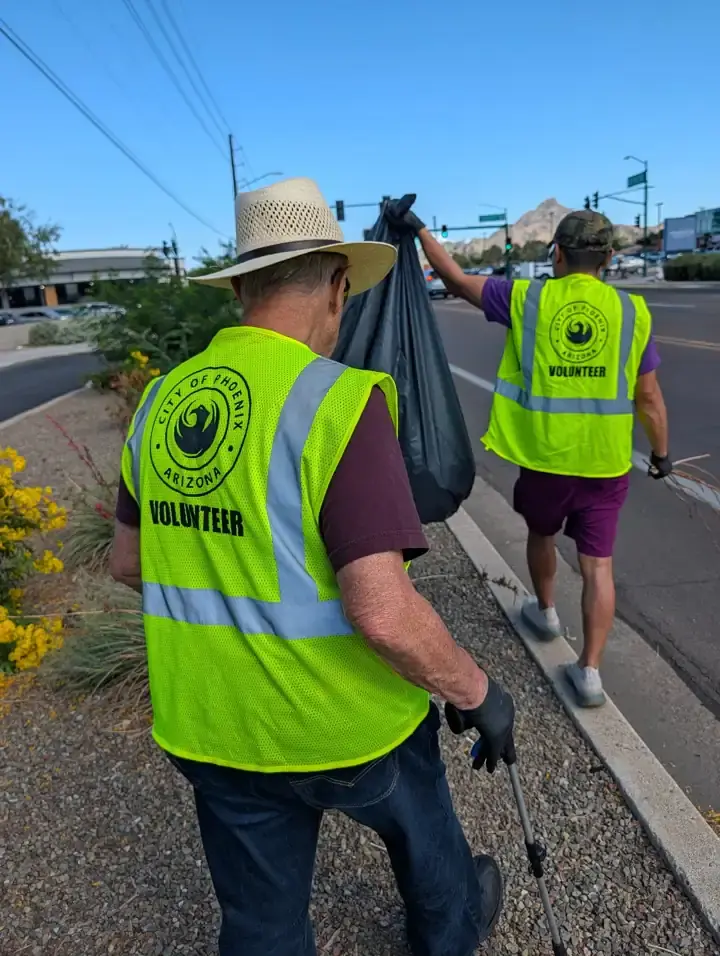 Volunteers wearing City of Phoenix safety vests carrying a filled trash bag beside the road