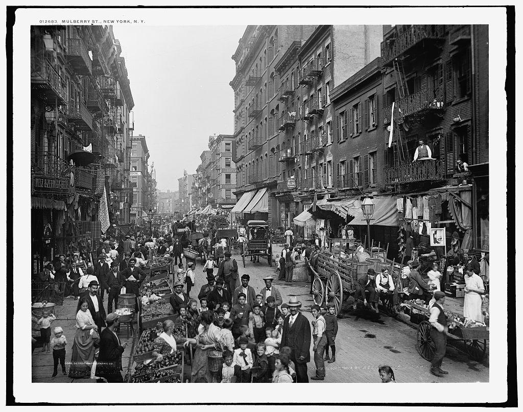 A historical view along Mulberry Street in New York, with pedestrians, market carts and buildings.