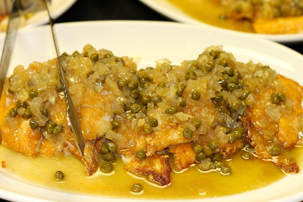 A platter of browned fish fillets in sauce with capers and serving tongs.