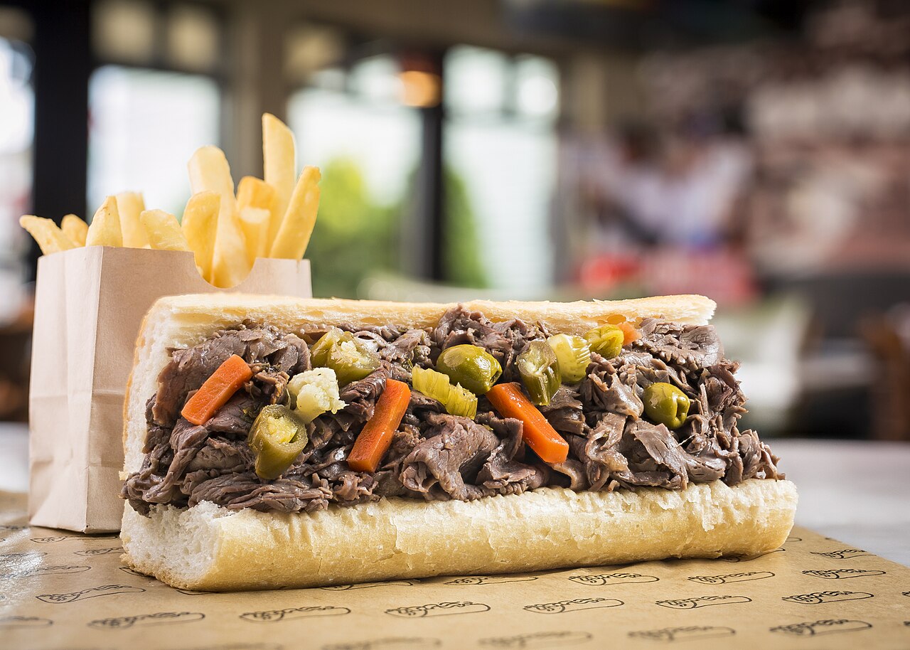 An Italian beef sandwich piled with sliced beef and colorful giardiniera, with fries behind it.