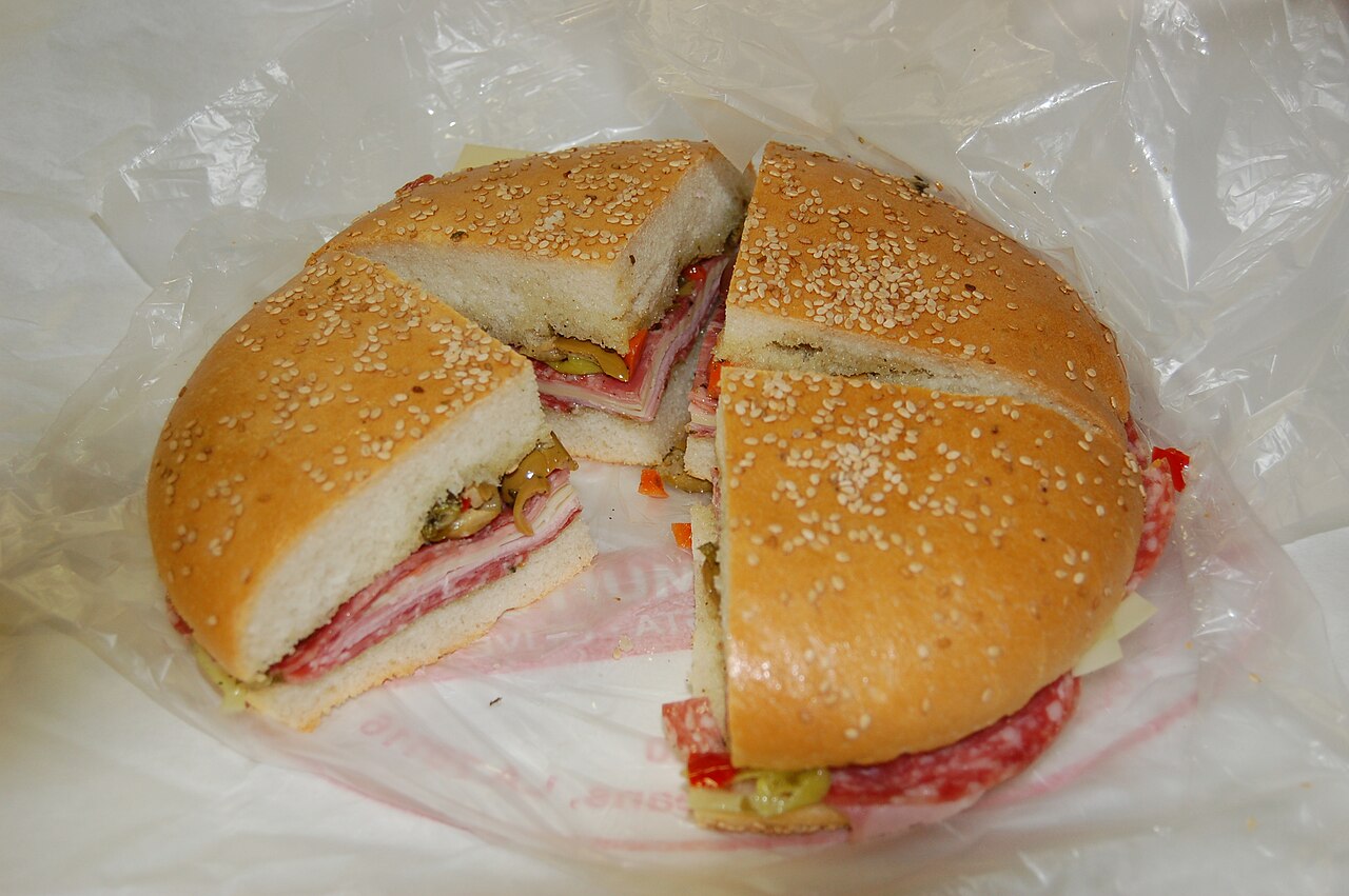 A round sesame loaf cut into four muffuletta quarters, showing layers of meat, cheese, and olive salad.