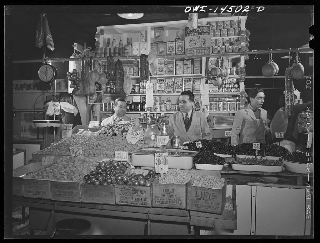 Italian American food shop in New York, with three men behind a counter of bulk foods and shelves of cans and jars.