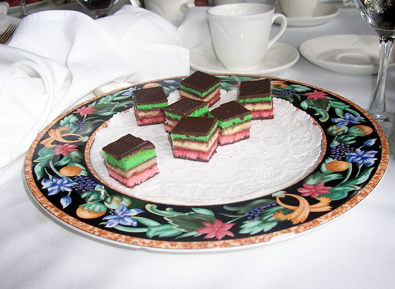 Chocolate-topped rainbow cookies with green, cream, and pink layers arranged on a patterned platter.