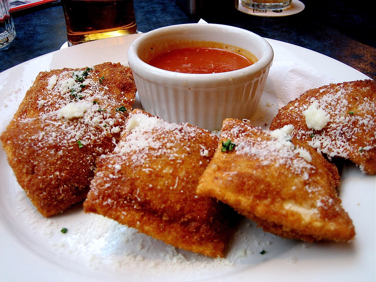 Golden toasted ravioli sprinkled with grated cheese and herbs, beside a ramekin of red dipping sauce.