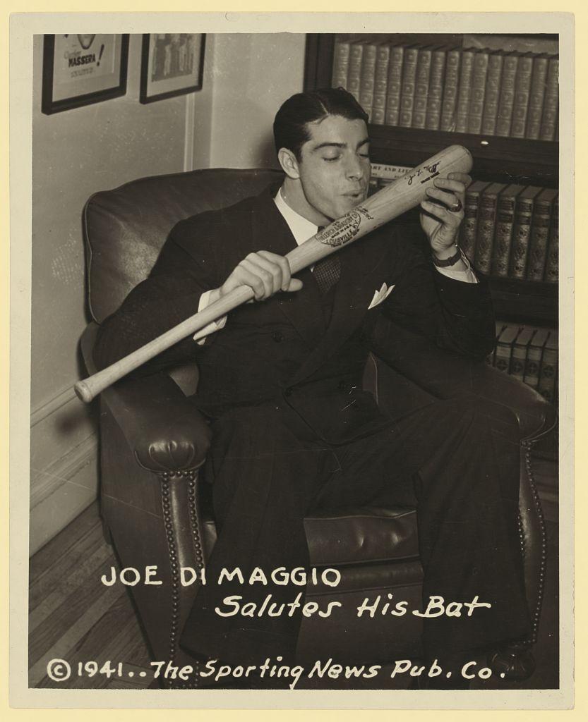 Joe DiMaggio seated in a suit, holding a baseball bat close to his face.